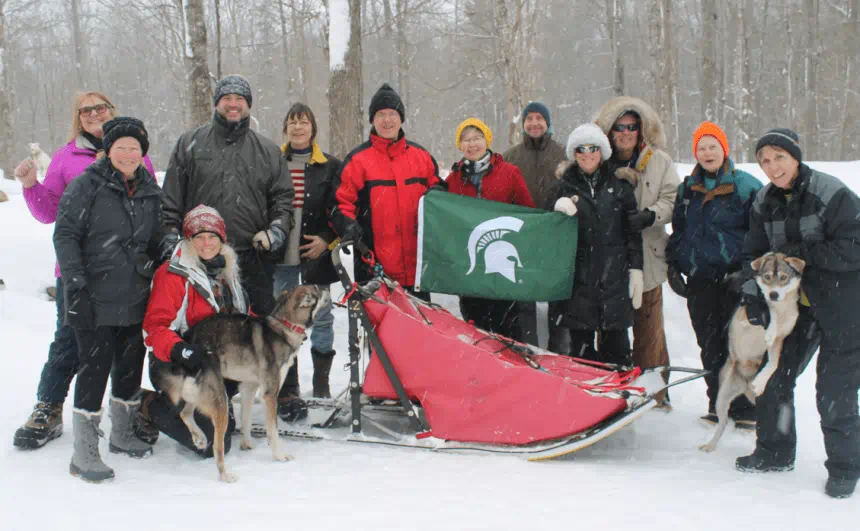 Group of people pose in a snowy setting with two sled dogs, a sled and a Spartan flag