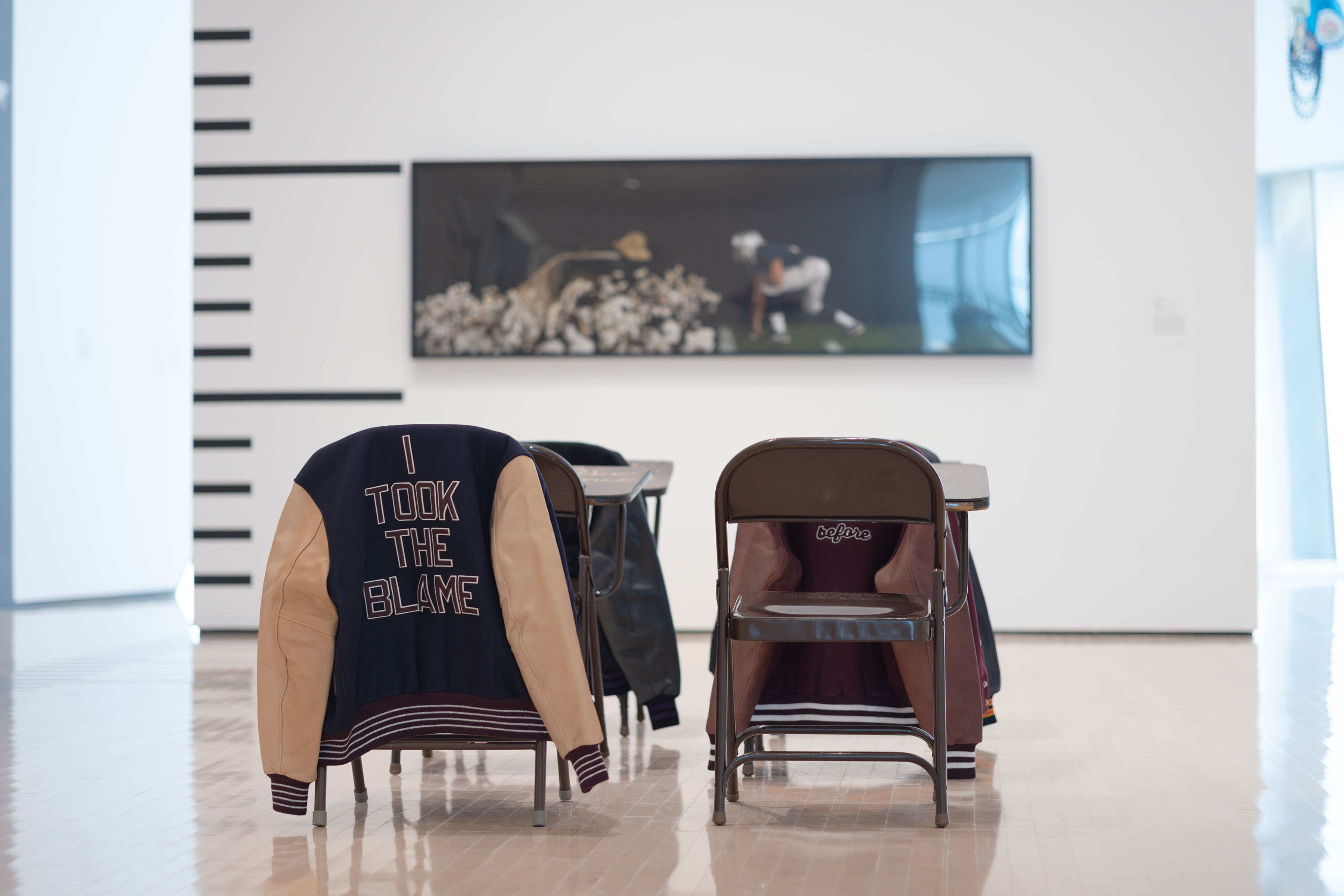  Classroom desks with varsity jackets hanging off the back of the chair in the MSU Broad ARt Museum’s “Resistance Training” exhibition. One of the jackets says, “I took the blame” on the back