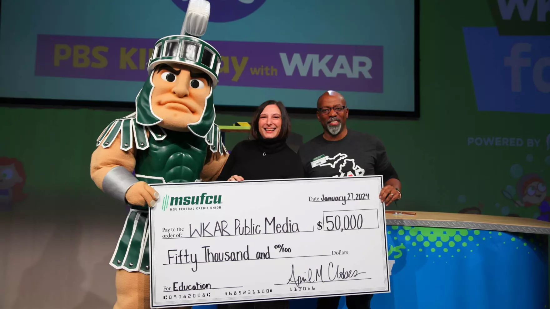 Sparty (left), April Clobes of MSU Federal Credit Union (middle) and Shawn Turner of WKAR (right) pose holding a large check from MSUFCU to WKAR Public Media for $50,000