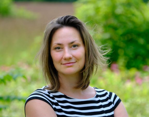 A portrait of a person with straight hair and a black-and-white striped shirt. In the background are green trees and foliage.