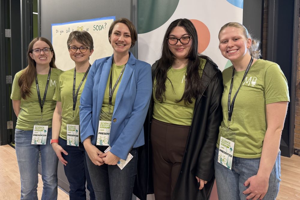 Five people stand in a row in front of a poster on a wall that says: “Do you call it pop or soda?” The people are wearing green shirts and lanyards with name tags that say: “MSU Science Festival Presenter.