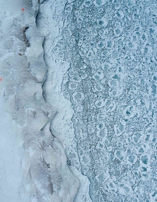 Aerial view of a textured icy landscape showing a natural pattern of frozen bubbles in a white snow-covered expanse, resembling a geological frost structure, highlighting the intricate details formed by winter conditions.