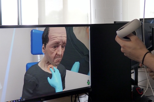  A hand controls a virtual reality avatar on a screen. The avatar is receiving an implant near its throat.