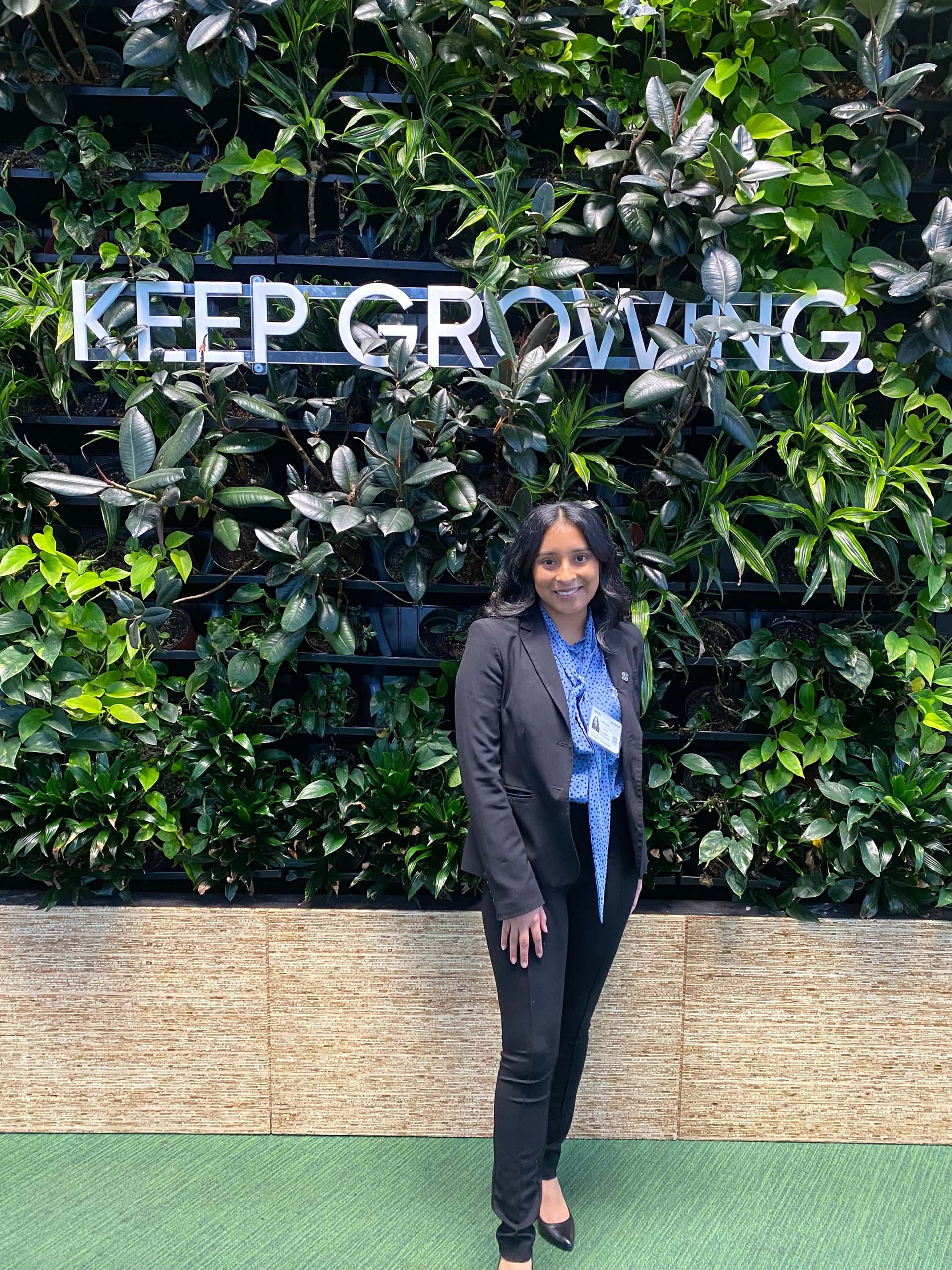 Anel Aguilar posing for a photo in front of a wall of plants with a sign that reads keep growing