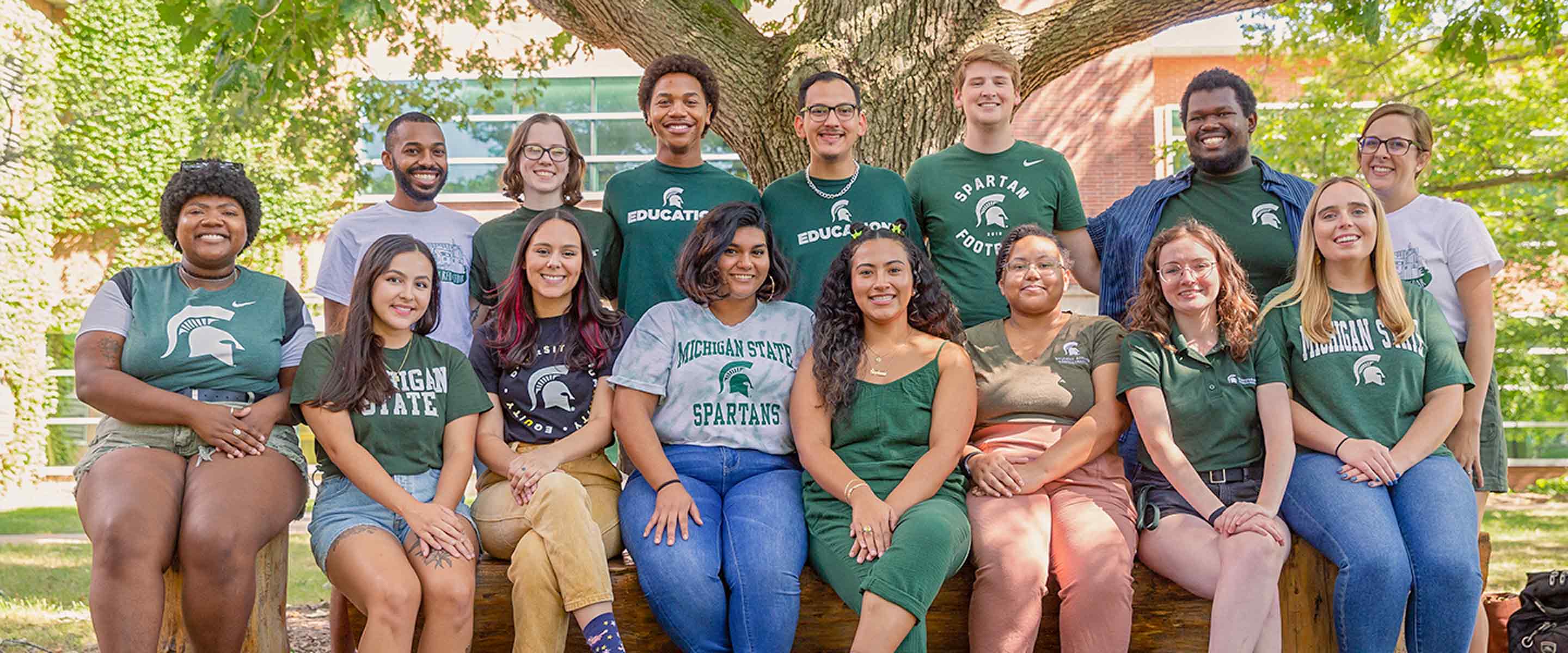 A diverse group of individuals is posed together, smiling, on a wooden bench structure outdoors. Many wear shirts in varying designs, all sporting the Michigan State Spartans logo, suggesting they are students or supporters of the institution. The group is framed by lush greenery and the trunk of a large tree, indicative of a campus setting. They appear to be relaxed and happy, conveying a sense of camaraderie and school spirit. The arrangement of the individuals, casual clothing, and the university insignia emphasize a collegiate atmosphere and possibly a student group or team.