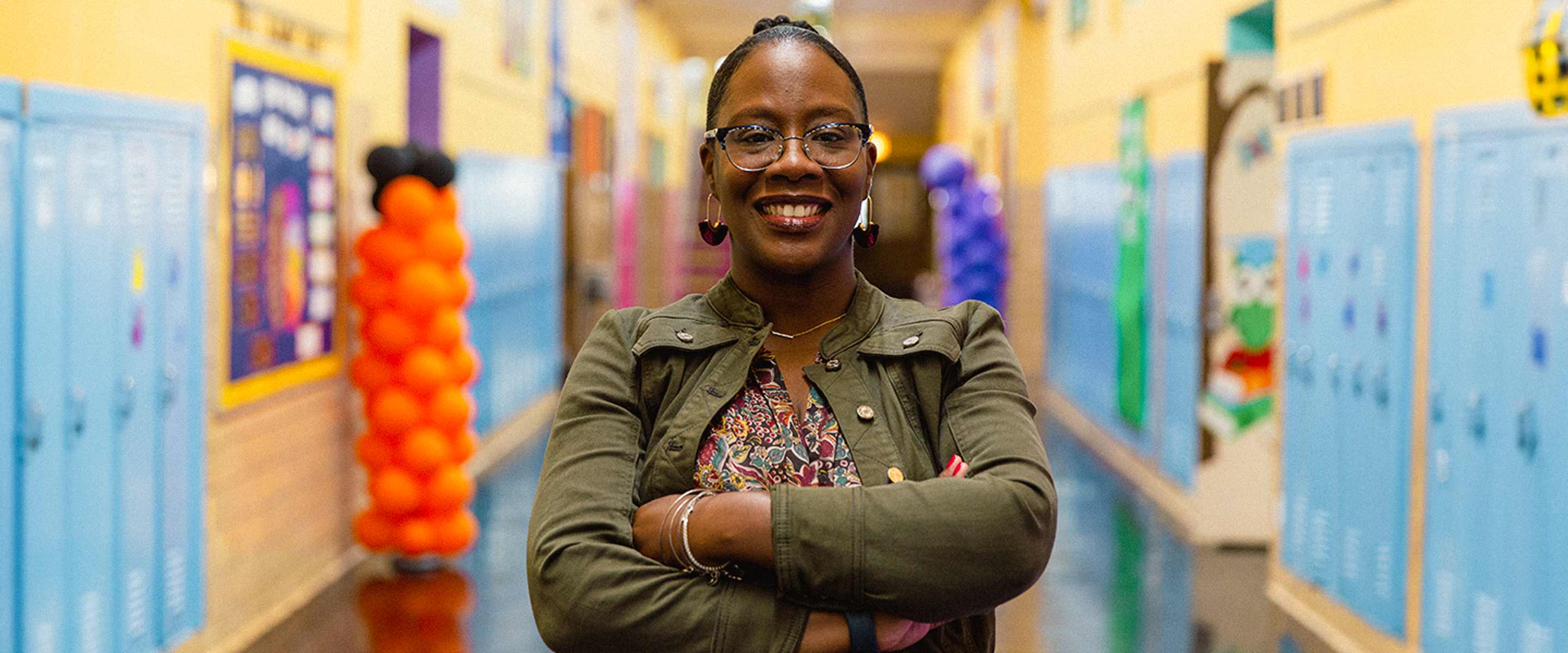 A person stands confidently with their arms crossed in the center of a vibrant school hallway, flanked by rows of lockers in varying shades of blue. They are wearing glasses, a patterned top, and a military green jacket, accessorized with hoop earrings and a pendant necklace. The background features colorful student lockers, educational posters, and a string of orange spherical decorations, suggesting a lively and dynamic educational environment. The focus on the individual, their attire, and the setting indicates their role may be significant within this school community.