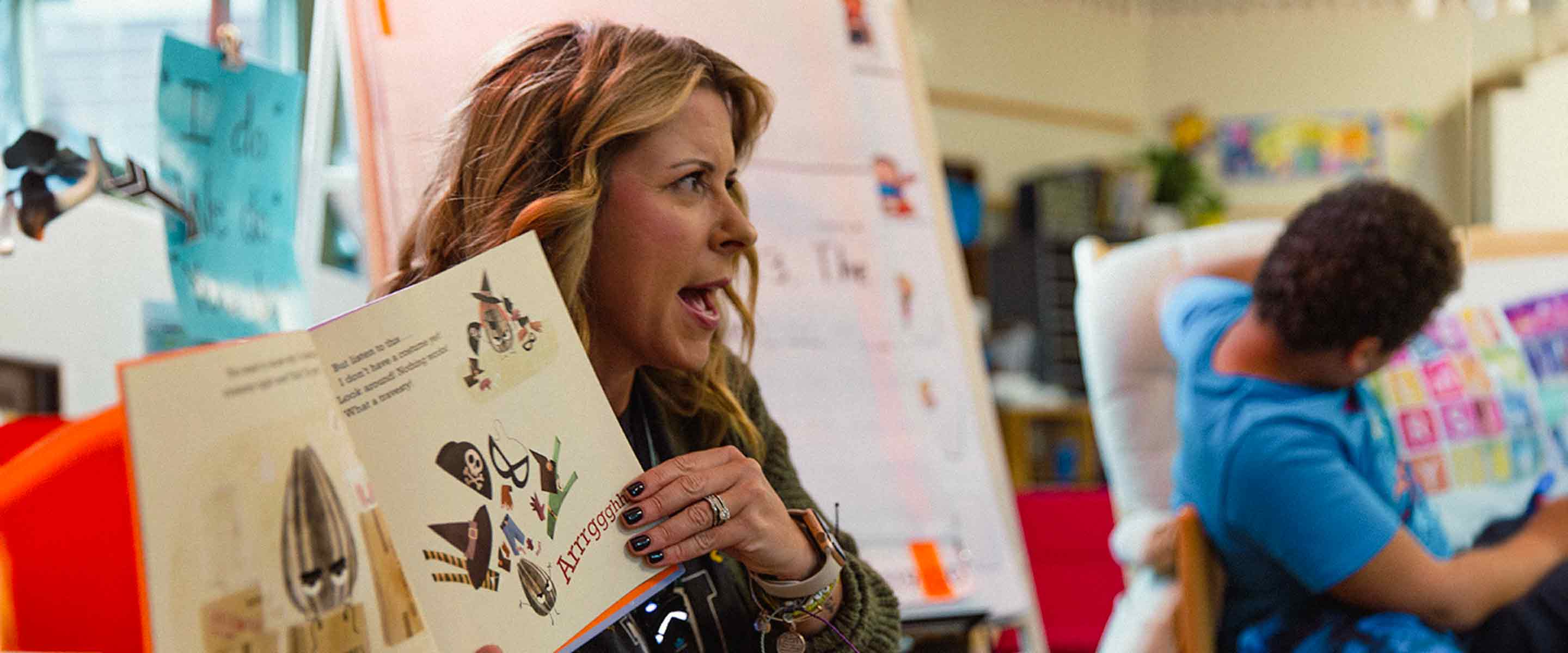 A person with shoulder-length hair and a wristwatch is energetically reading aloud from a book to an audience, not in the frame. They are wearing a dark cardigan and rings on their fingers. The book cover features playful illustrations, possibly related to children's literature. In the background, a large paper is mounted on the wall with various colorful visual aids, suggesting an educational or classroom setting. Another individual, partly out of focus and facing away from the camera, is seated nearby, engrossed in reading or studying a document with a vibrant pattern.