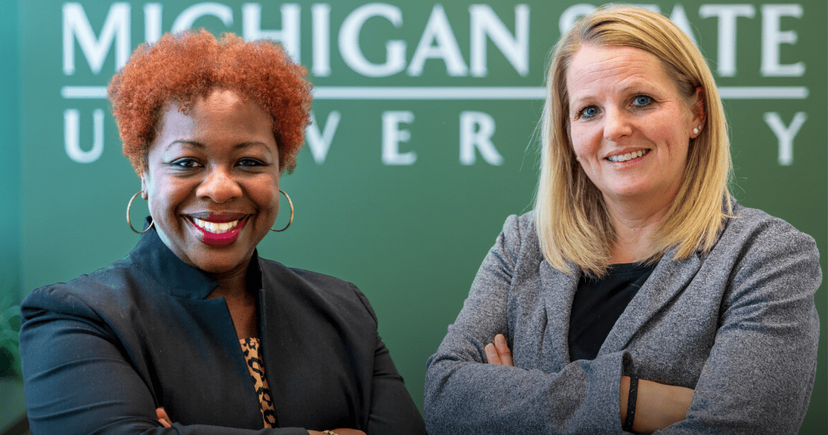 Sheneka Williams (left) and Emily Bouck (right) stand in front of the green backdrop of Michigan State University.