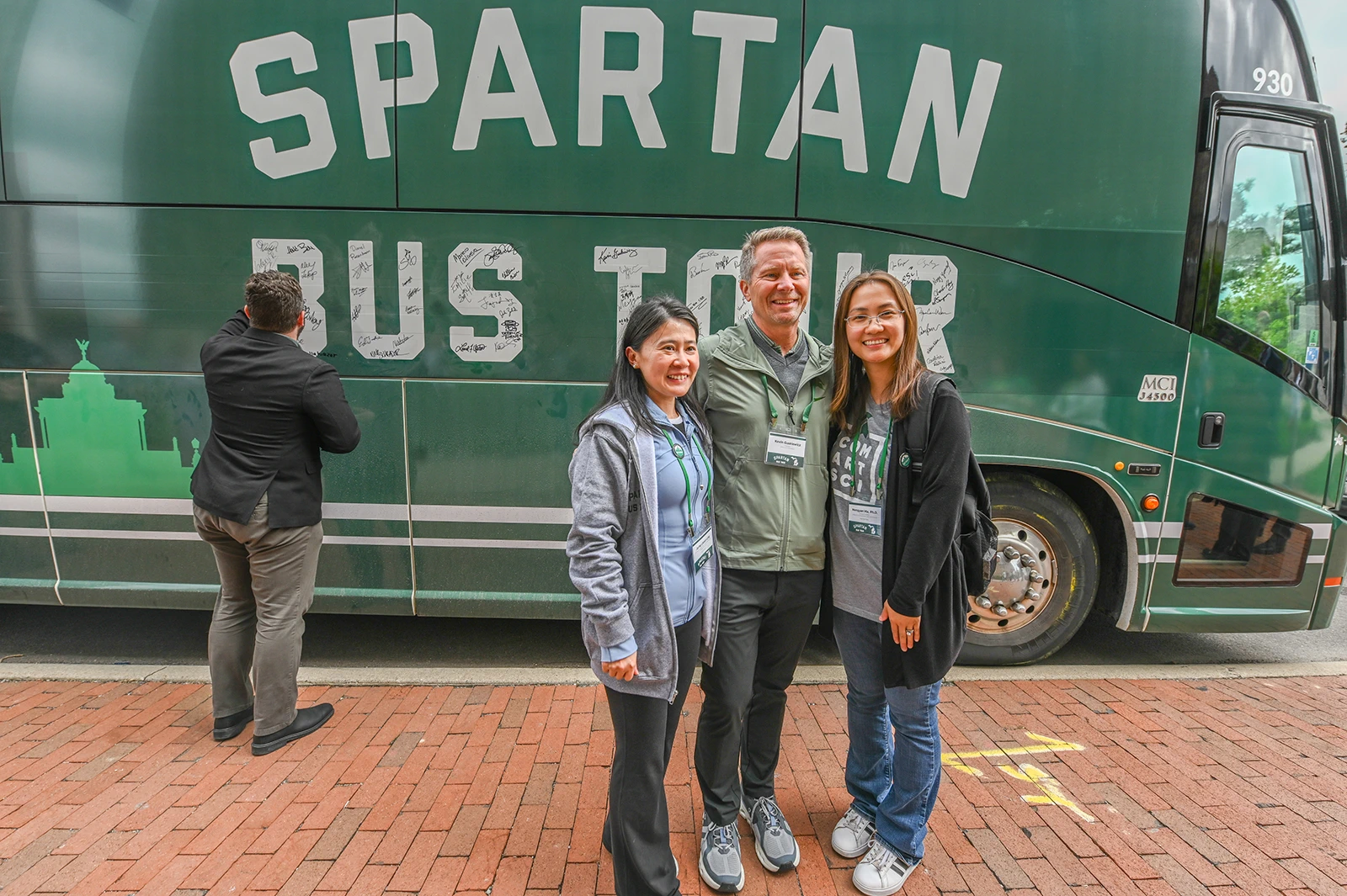 Three people pose for a photo in front of a bus. The bus is green and “Spartan Bus Tour” is printed on it in bold white letters.
