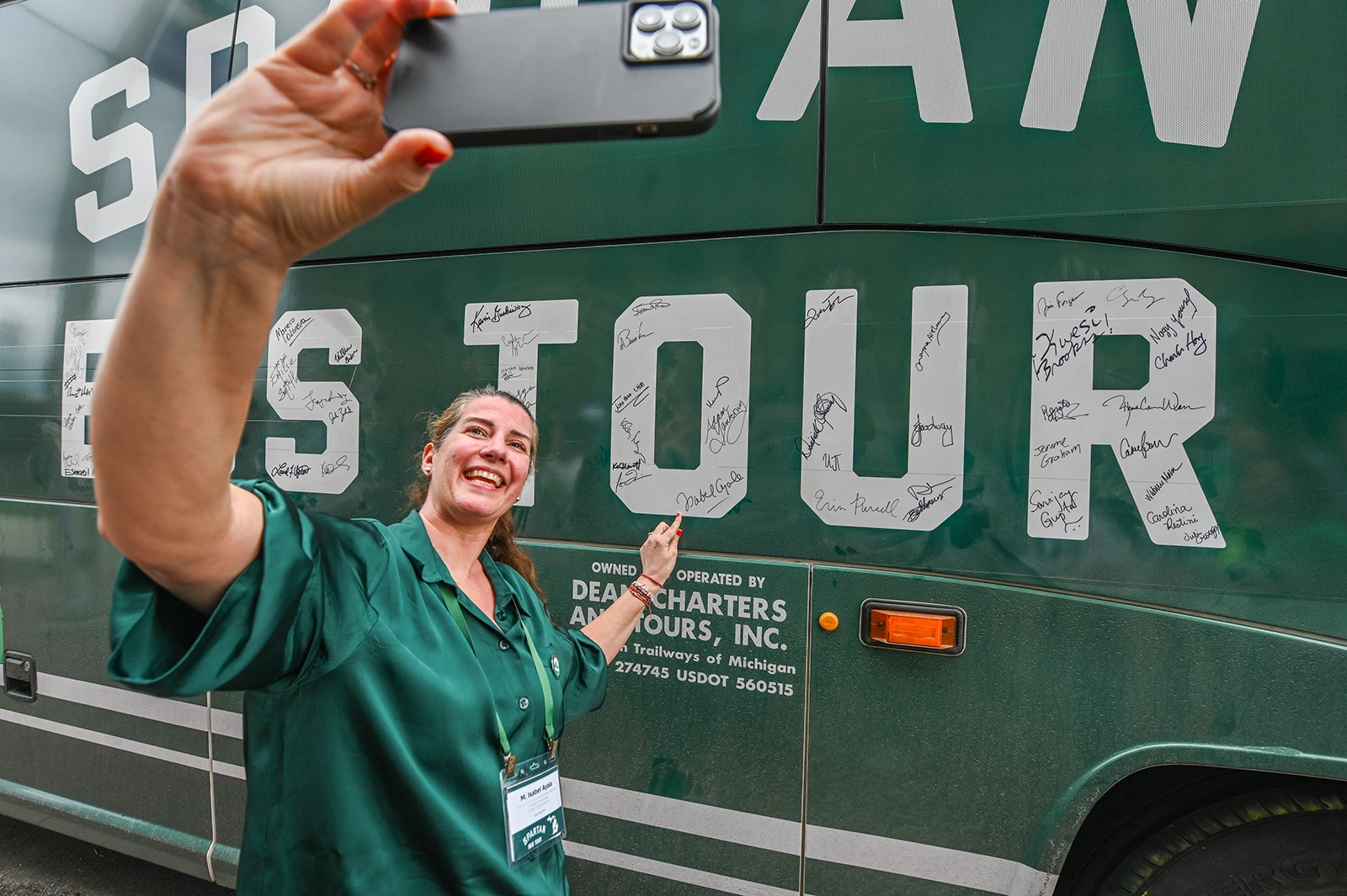 A woman taking a selfie with a bus that says “Spartan Bus Tour” in white block letters. There are signatures inside the block letters. She is pointing to a signature and smiling.