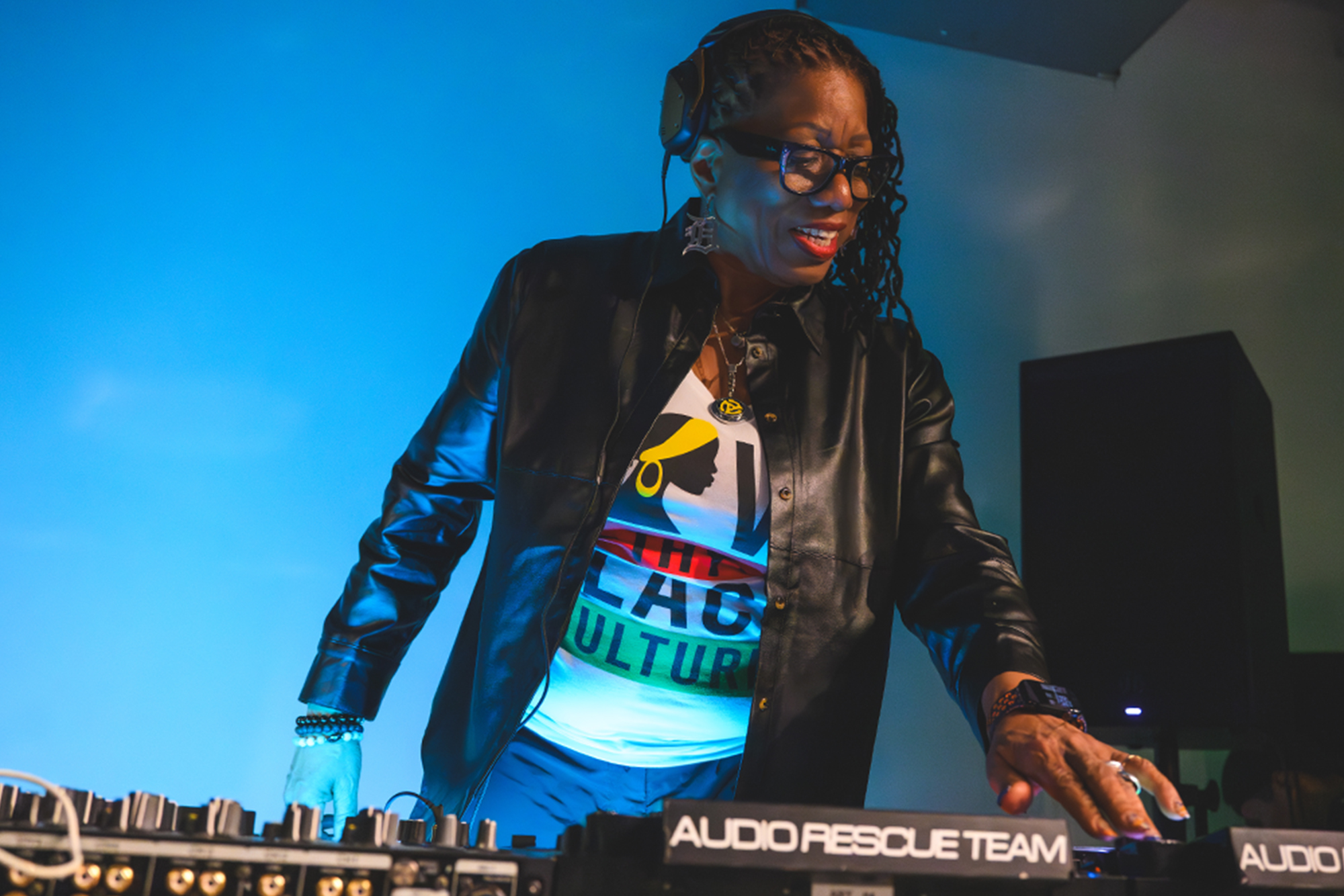 DJ Stacey “Hotwaxx” Hale wearing a black leather jacket and headphones mixes music behind a sound mixing board