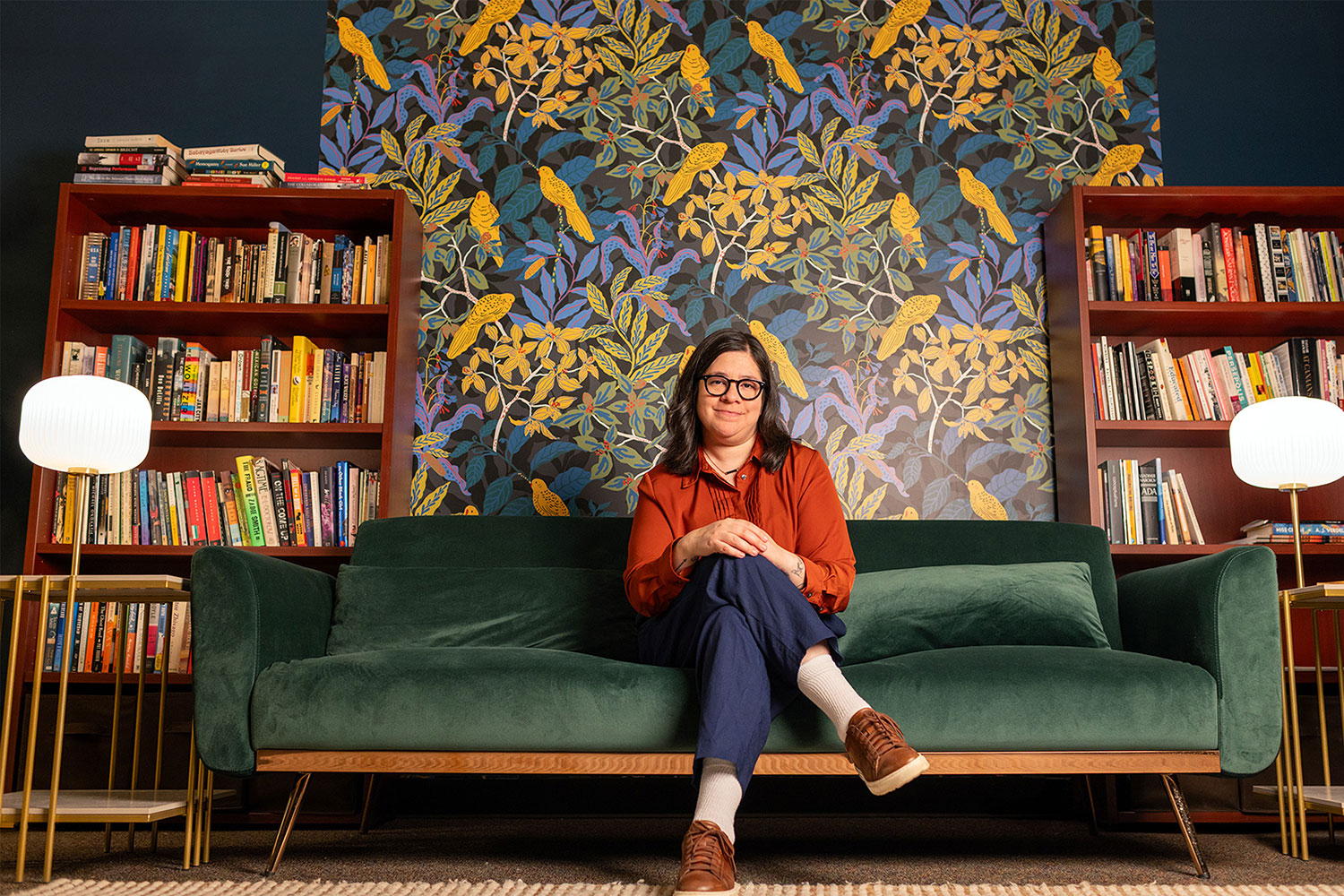Alejandra Márquez, assistant professor of Spanish at Michigan State University, wears an orange shirt and glasses. She sits in a yellow chair and reads a book. A floor lamp is on the left side of the chair. A large indoor plant is on the right side of the chair.
