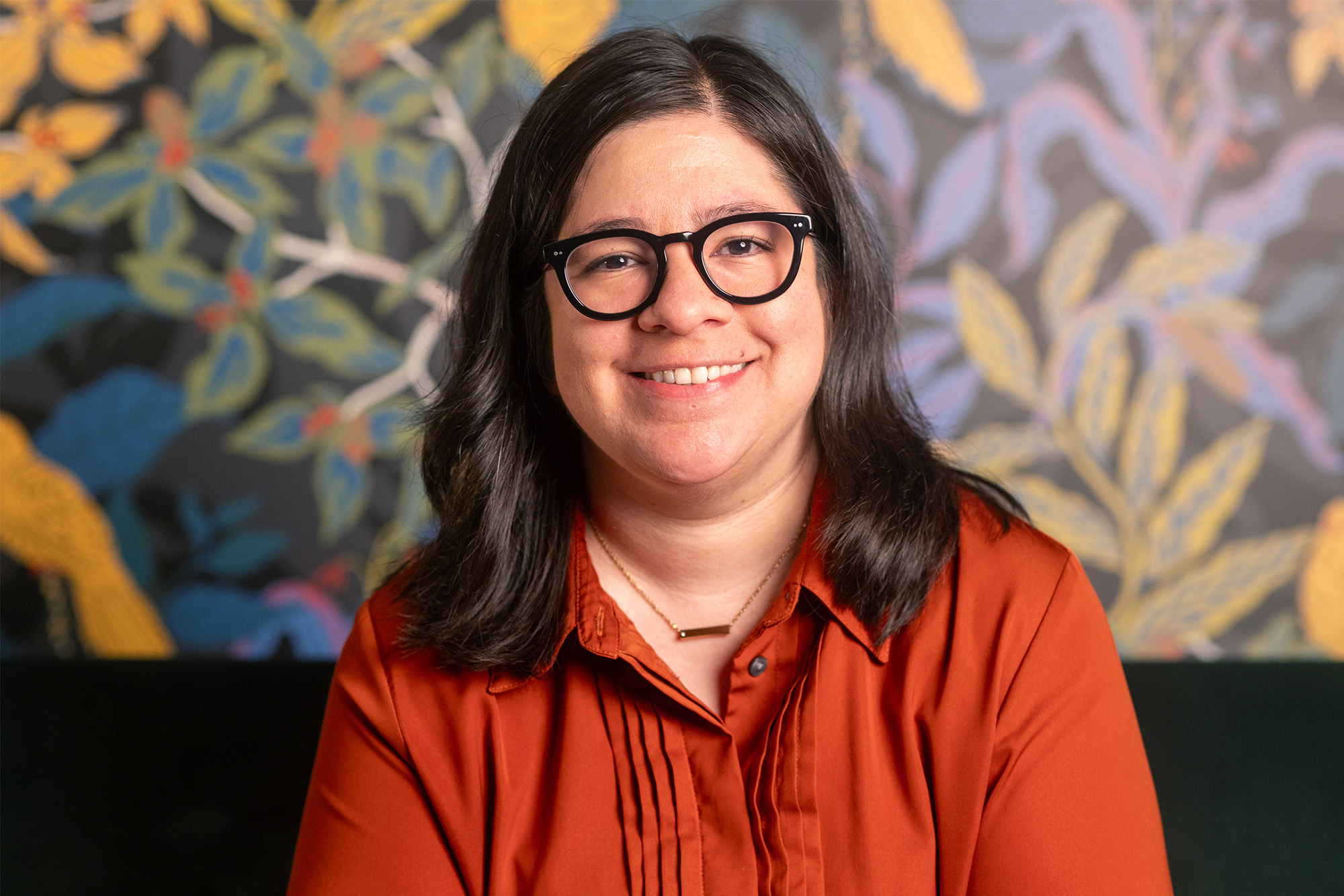 Alejandra Márquez, assistant professor of Spanish at Michigan State University, wears an orange shirt and glasses. She sits in from a wall with wallpaper that shows green, blue, and yellows leaves.