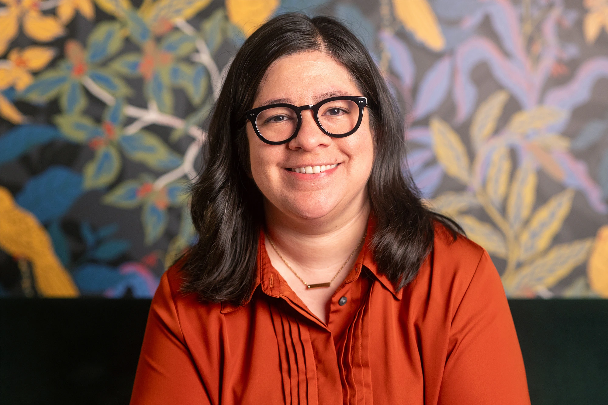 Alejandra Márquez, assistant professor of Spanish at Michigan State University, wears an orange shirt and glasses. She sits in from a wall with wallpaper that shows green, blue, and yellows leaves.