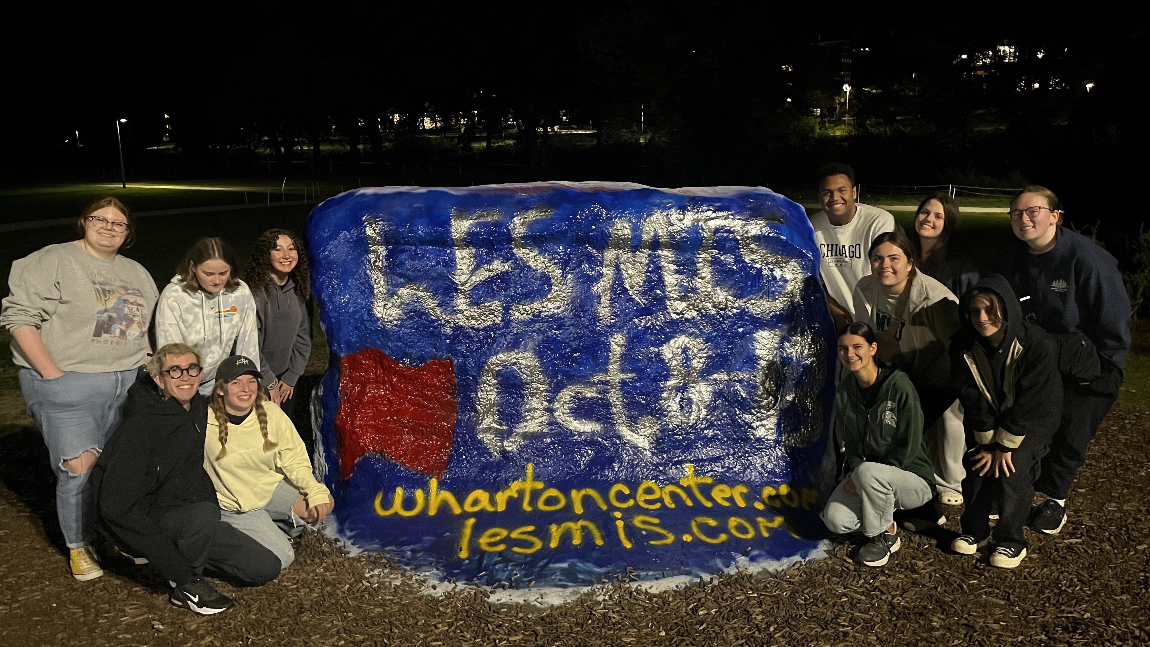 A group of people around the Rock painted blue with "Les Mes Oct. 8" and a URL to Wharton Center on top.