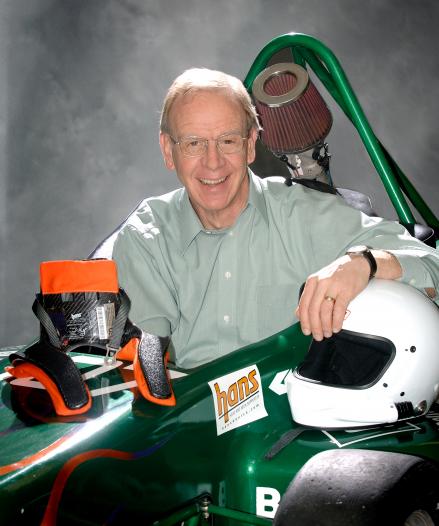 A man smiles in a green shirt while sitting in a race car. He has his left hand atop a driving helmet and his right atop a HANS, or head and neck support, device. 