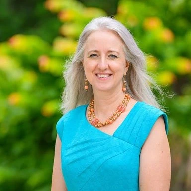 Portrait of Stephanie Nawyn, a smiling woman with long gray hair, wearing a turquoise dress and orange beaded jewelry, standing in front of green foliage.