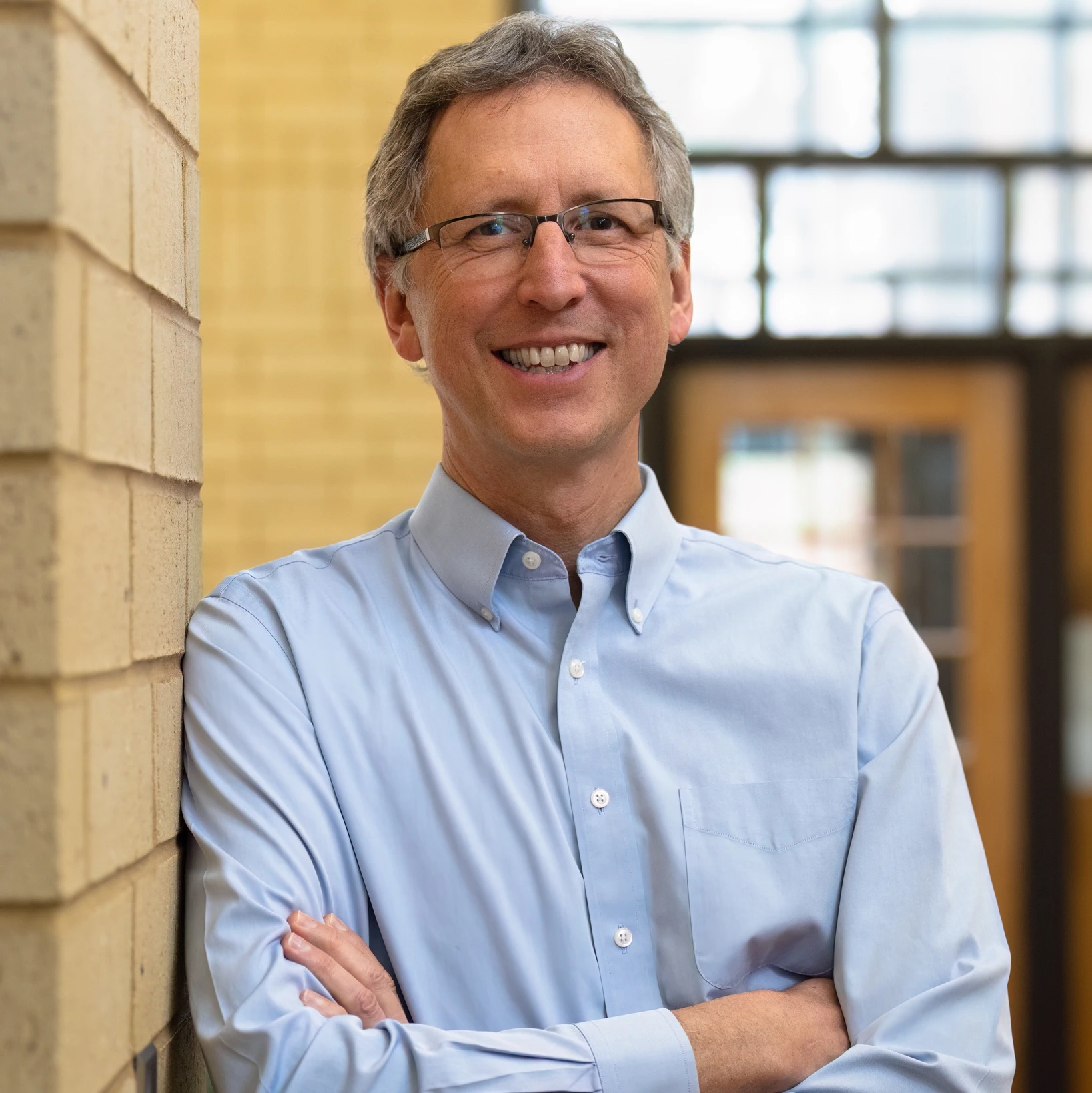 A man wearing glasses smiles while leaning against a brick wall and crossing his arms