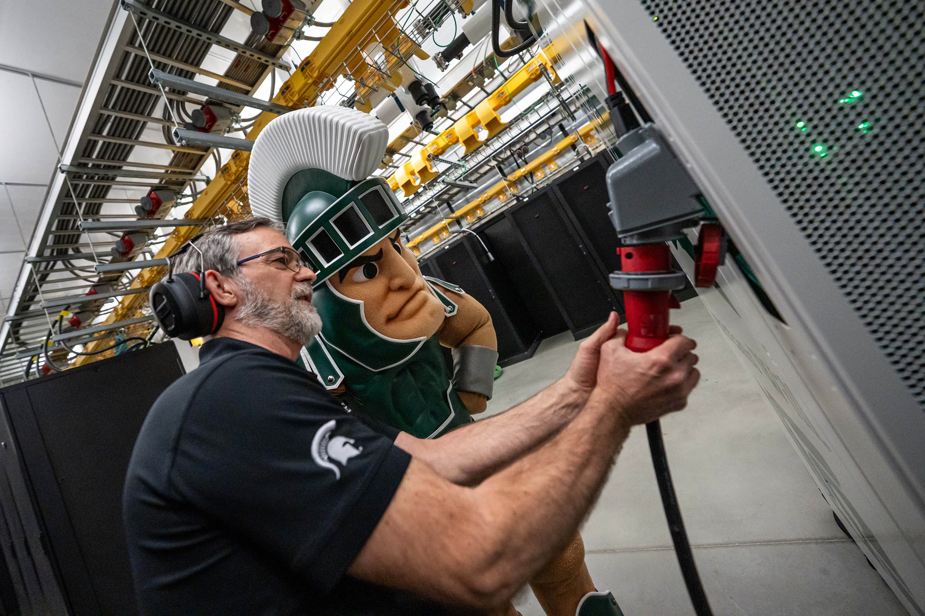 A system administrator inserts a large red plug into a gray socket in an industrial room while MSU’s Sparty mascot observes.