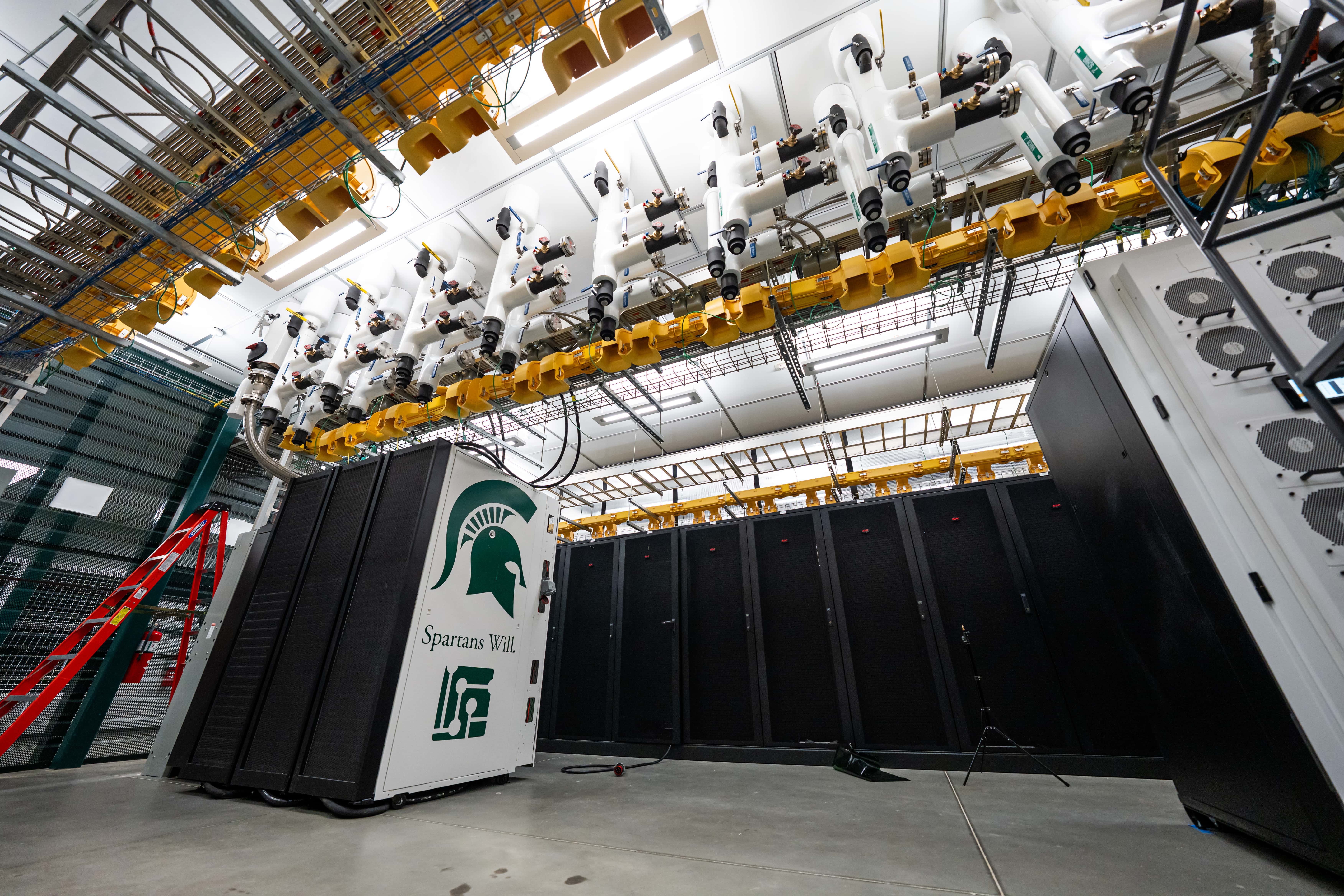 A large industrial room filled with tall racks of computer hardware, wires, and pipes projecting from the ceiling. One of the racks is decorated with a green Spartan helmet, the words
