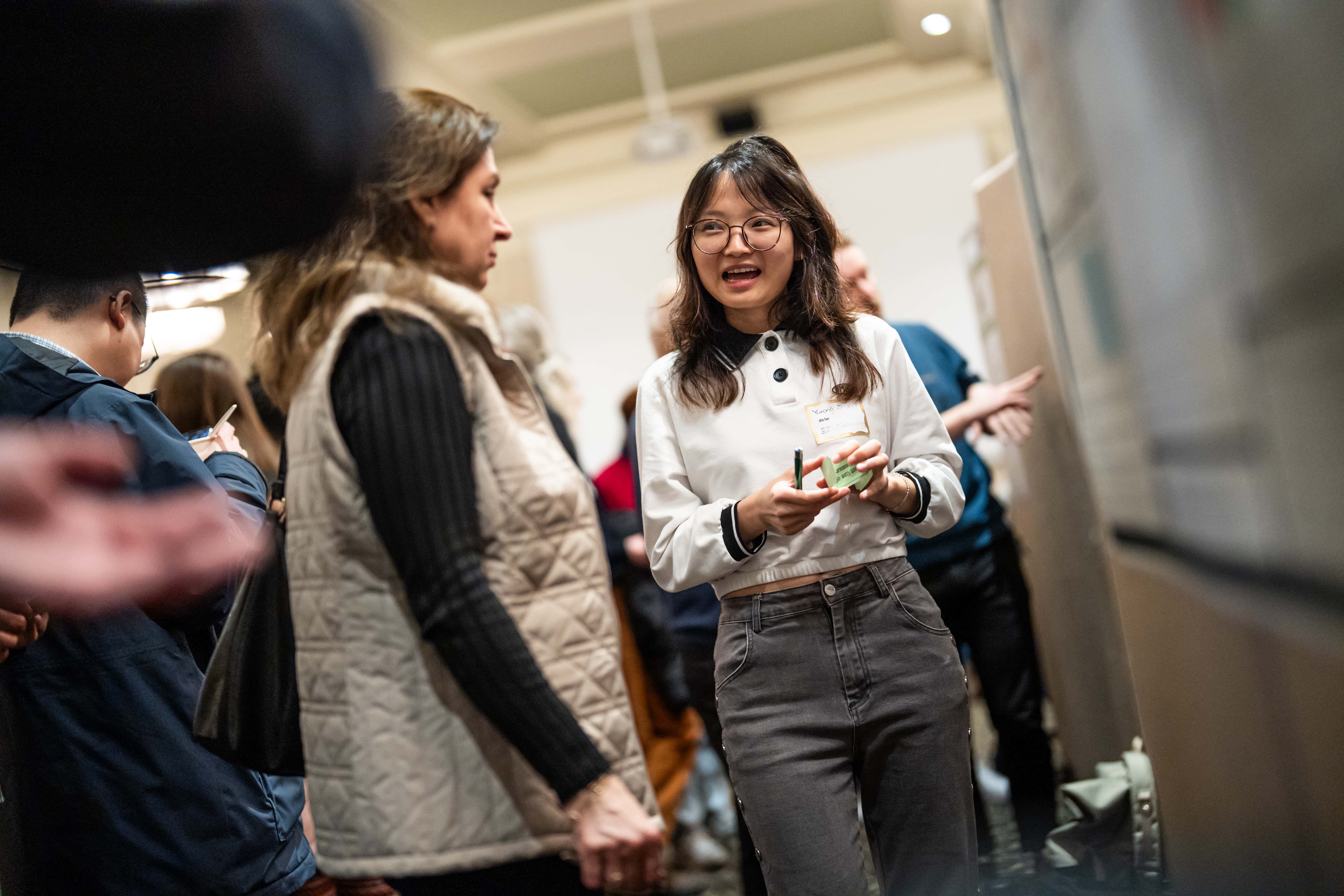 People gather in a room to view academic posters. A student in focus at the center of the image presents to an attentive visitor.