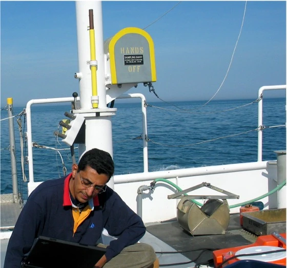 A person works on a laptop aboard a research vessel on a Great Lake. The individual is seated near scientific equipment, including a sediment grab sampler and tubing. A yellow sign on a white post reads