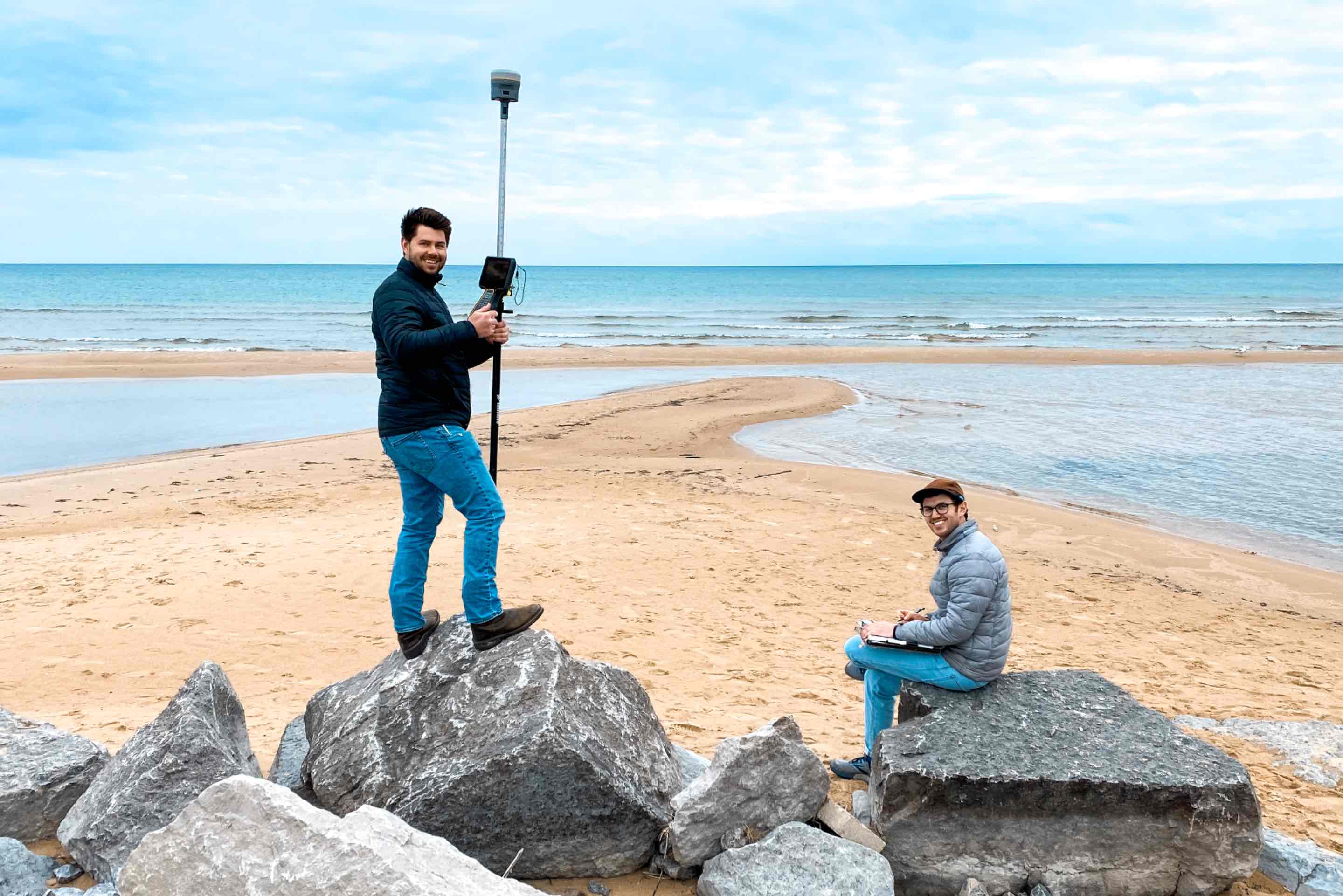 Two people conduct fieldwork on a rocky shoreline. One person stands on a large rock holding a GPS surveying instrument with a mounted tablet, while the other sits nearby on a rock writing on a clipboard. A sandy beach, calm water, and cloudy sky are visible in the background.